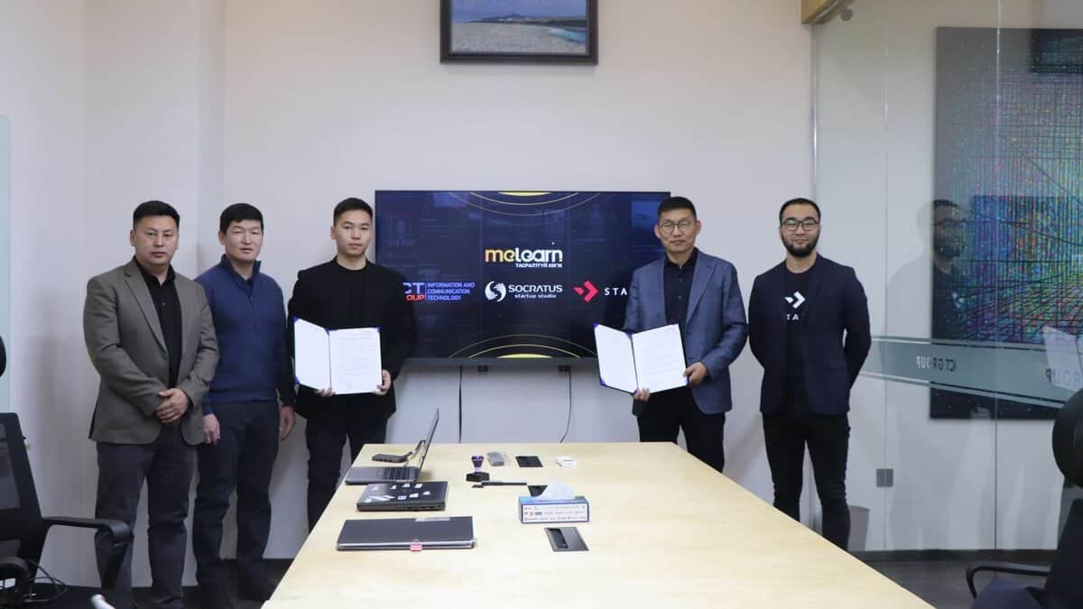 Зургаан хүн хурлын танхимд зогсож, хоёр баримт бичиг барьж байна. Тэдний ард олон логог харуулсан дэлгэц байдаг. Урд талын ширээн дээр янз бүрийн электрон төхөөрөмжүүд байдаг. - Business.MN