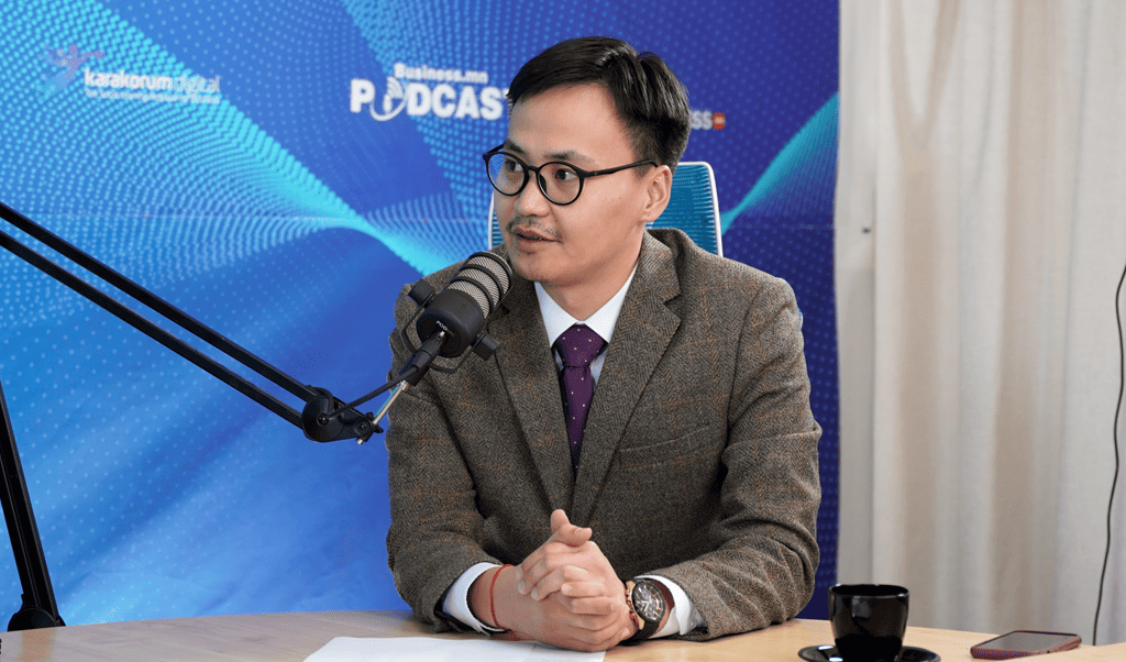 Нүдний шил, бор пиджак, нил ягаан зангиа зүүсэн хүн урд микрофонтой ширээний ард сууна. Тэд подкаст бичлэг хийж эсвэл оролцож байгаа бололтой. Арын дэвсгэр дээр "Business Podcast" гэсэн үгс болон энтрепренерийн сэдэвтэй холбоотой зарим график элементүүд харагдана. - Business.MN