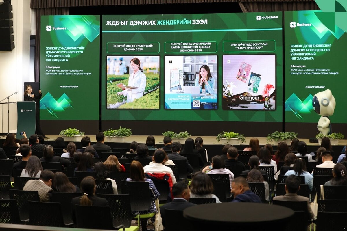 An audience sits facing a stage with large screens displaying business presentations and images of entrepreneurs. The event appears professional, with a presenter at a podium and plants decorating the stage.