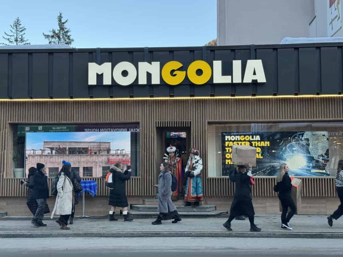 People walk past a building with a "MONGOLIA" sign. Traditional Mongolian mannequins are displayed at the entrance. The scene includes a large screen displaying various images.
