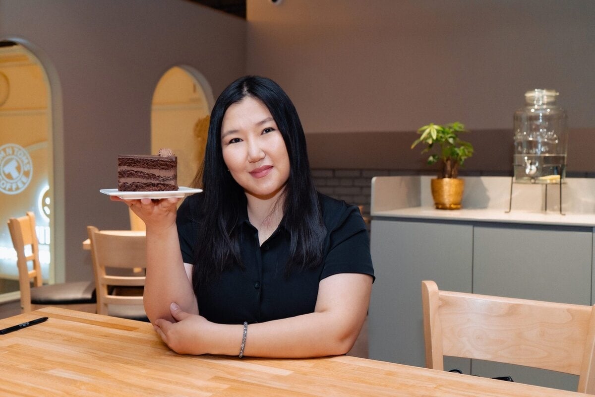 Урт хар үстэй, хар цамц өмссөн эмэгтэй кафед модон ширээний ард инээмсэглэн сууж, шоколадтай бялууны зүсмэл бүхий таваг өргөж байна. Арын дэвсгэр дээр цайвар өнгийн сандал, ваартай ургамал байдаг. - Business.MN