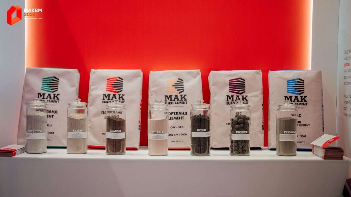 Цаана нь "МАК Евро цемент" гэсэн шошготой ууттай шилэн саванд хийсэн янз бүрийн төрлийн цемент. Лааз бүр өөр өөр төрлийн цемент агуулсан бөгөөд хажуу талд нь мэдээллийн товхимол байдаг. Улаан, цагаан дэвсгэр нь дэлгэцийг онцолж, ард байгаа компанийн бизнесийн ур чадварыг харуулдаг. - Business.MN