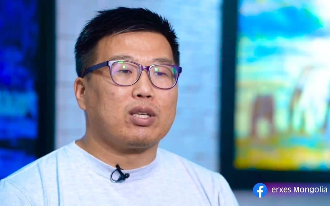 Богино хар үстэй, нүдний шил, цайвар саарал цамц өмссөн эрэгтэй хөх, ногоон өнгийн бүдгэрсэн дэвсгэрийн өмнө ярьж байна. Түүний цамцан дээр микрофон хавсаргасан байдаг. Баруун доод буланд Facebook-ийн лого "erxes Монголын бизнес компани. - Business.MN