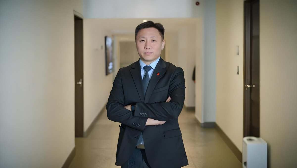 A man in a suit stands in a well-lit corridor with beige walls and closed doors. He has short hair and his arms are crossed. The hallway is empty aside from a white appliance on the floor.