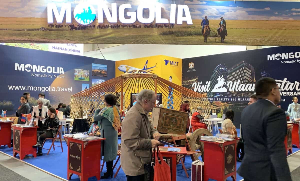 Монголын үзэсгэлэнт газруудыг харуулсан хөл хөдөлгөөн ихтэй худалдааны лангуу. Оролцогчид уламжлалт юртыг оролцуулан үзэсгэлэнг үзэж сонирхдог. Баннер, зурагт хуудас нь аялал жуулчлал, соёлын туршлагыг онцлон тэмдэглэж, хүмүүс ширээний ард сууж байна. - Business.MN