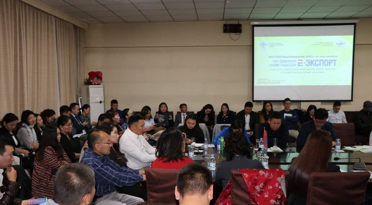 Төрөл бүрийн бүлэг хүмүүс хурлын танхимд сууж, танилцуулга үзүүлж буй проекторын дэлгэц рүү анхааралтай хандана. Өрөө нь гэрэлтүүлэг сайтай, оролцогчид оролцож байгаа нь албан ёсны уулзалт эсвэл семинар зохион байгуулахыг санал болгож байна. - Business.MN