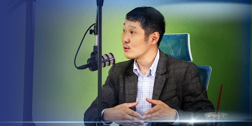 Богино хар үстэй хүн ногоон дэвсгэрийн өмнө ширээний ард сууж, индэр дээр микрофоноор ярьж байна. Тэд энтрепренерийн дүр төрхийг илэрхийлсэн эрээн цамц, бараан пиджак өмссөн байна. Ширээн дээр улаан сүрэлтэй ундаа байна. Зураг нь цэнхэр өнгийн градиент давхаргатай. - Business.MN