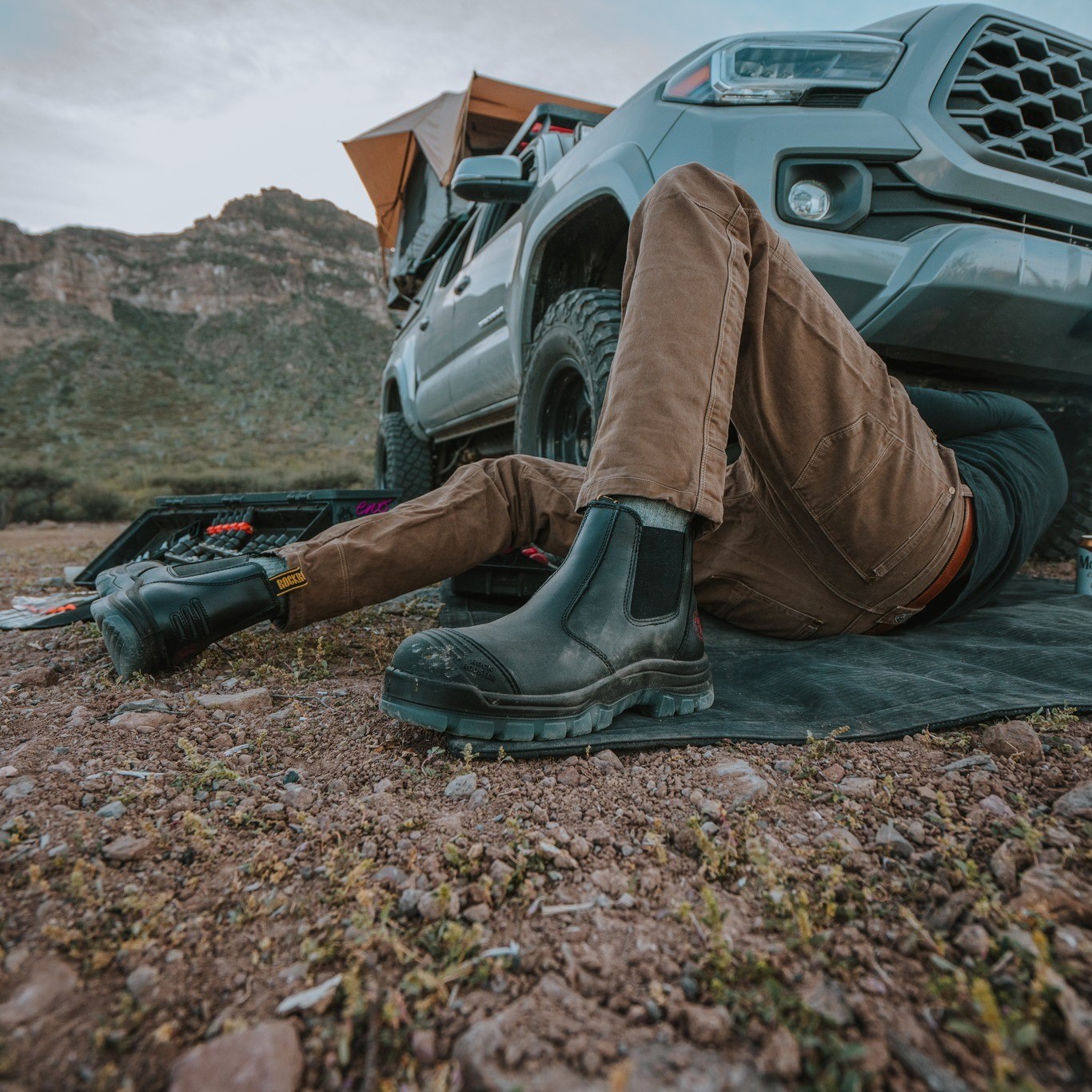 A person wearing brown pants and black boots lies on a mat under a parked off-road vehicle, possibly working on it, with tools scattered nearby in a rugged, mountainous outdoor setting.