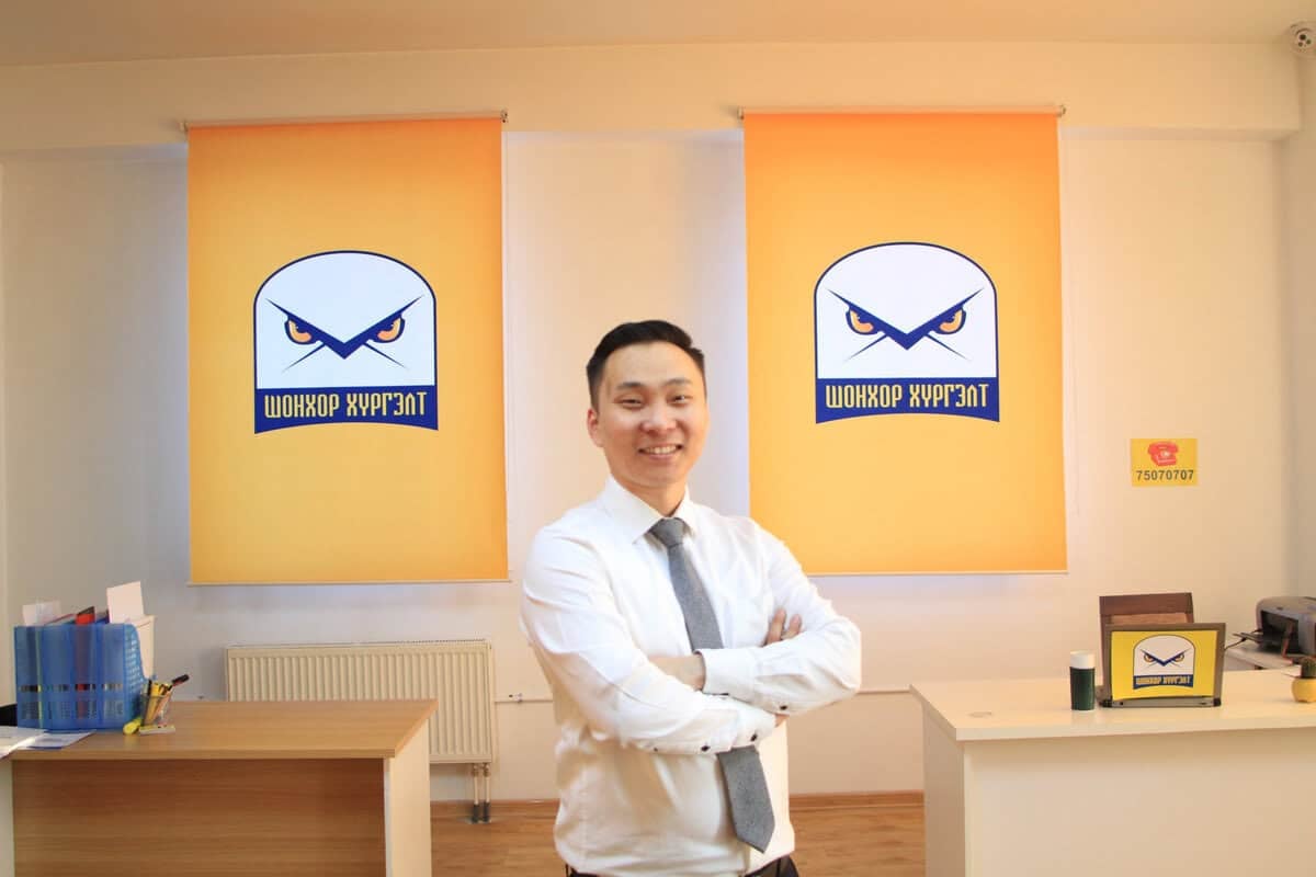 Цагаан цамц, зангиа зүүсэн эр албан тасалгаанд гараа зөрүүлэн инээмсэглэн зогсож байна. Түүний ард бүргэдийн дүрс бүхий лого бүхий хоёр туг байна. Албан тасалгааны хэрэгслүүдтэй ширээ урд талд харагдана. - Business.MN