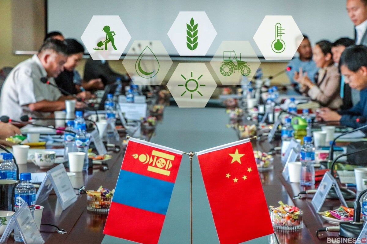 Ширээн дээр Монгол, Хятадын төрийн далбаатай уулзалт үргэлжилж байна. Дээрх төрөл бүрийн дижитал дүрс нь хөдөө аж ахуй, температур, ус, нарны гэрлийг илэрхийлдэг. Оролцогчид микрофон, баримт бичиг бүхий том хурлын ширээний эргэн тойронд сууж байна. - Business.MN