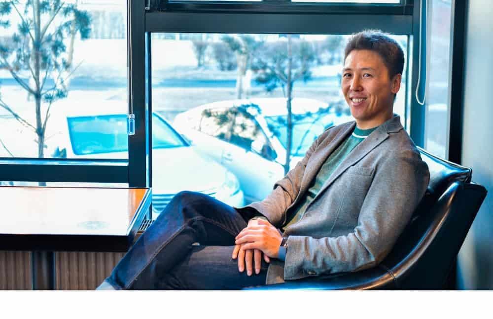 Бизнес-энтрепренер цонхны дэргэдэх хар сандал дээр тухтай сууж, камер руу инээмсэглэв. Тэрээр ногоон цамц, цэнхэр жинсэн өмд дээр саарал пиджак өмссөн байна. Цонхны гадна талд зогссон машинууд, цастай моднууд харагдана. - Business.MN