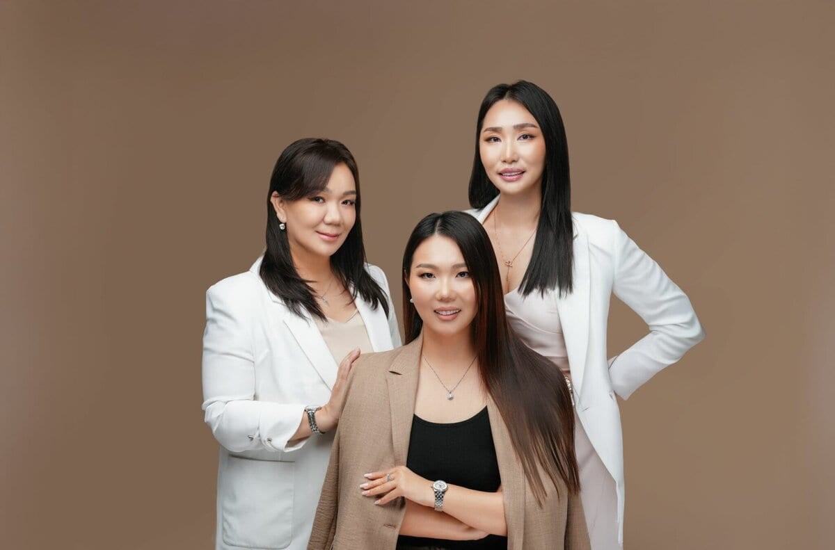 Тэнцвэртэй амьдралыг шингээсэн гурван эмэгтэй энгийн бор дэвсгэр дээр хамтдаа зургаа авахуулж байна. Хоёр нь цагаан пиджактай, гурав дахь нь хар топ дээгүүр бор шаргал өнгөтэй пиджактай урд сууж байна. Бүгд инээмсэглэн камер руу харж байна. - Business.MN