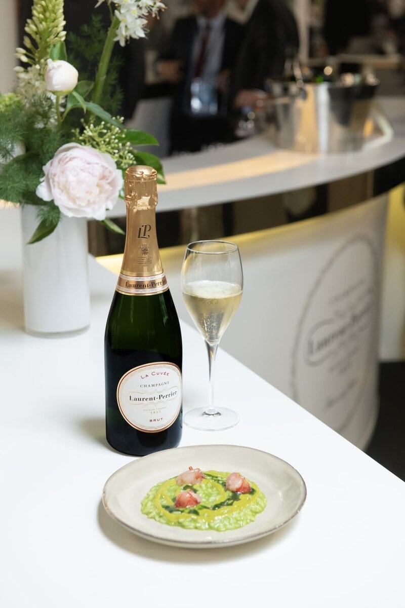 A bottle of Laurent-Perrier champagne and a filled glass sit on a white counter next to a dish of green risotto garnished with tomatoes. There's a floral arrangement nearby, and a bar area with people in the background.