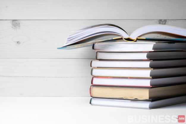 Модон дэвсгэртэй овоолсон ном. Дээд талын ном нээлттэй, яндан дээр хэвтэж байна. Business.mn лого баруун доод буланд харагдана. - Business.MN