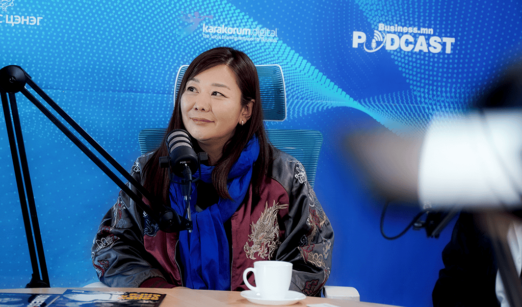 Урт хар үстэй эмэгтэй подкаст студид сууж байхдаа микрофоноор ярьж байна. Тэрээр нарийн хийцтэй хар хүрэм, цэнхэр ороолт өмссөн байна. Түүний урд ширээн дээр цагаан аяга сууна. Арын дэвсгэр дээр "Business.mn PODCAST" гэсэн бичвэр гарч байгаа бөгөөд түүнийг Karakorum Digital компанийн энтрепренер гэж онцолсон байна. - Business.MN