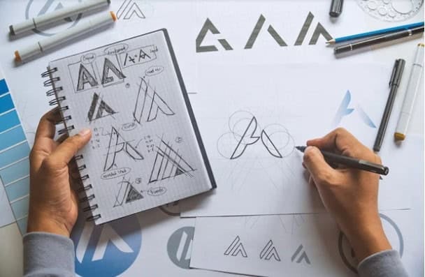 Өсөн нэмэгдэж буй компанийн лого дизайныг цаасан дээр болон дэвтэр дээрээ тэмдэглэгээ болон бусад зургийн хэрэгслээр хүрээлүүлсэн гарууд. - Business.MN