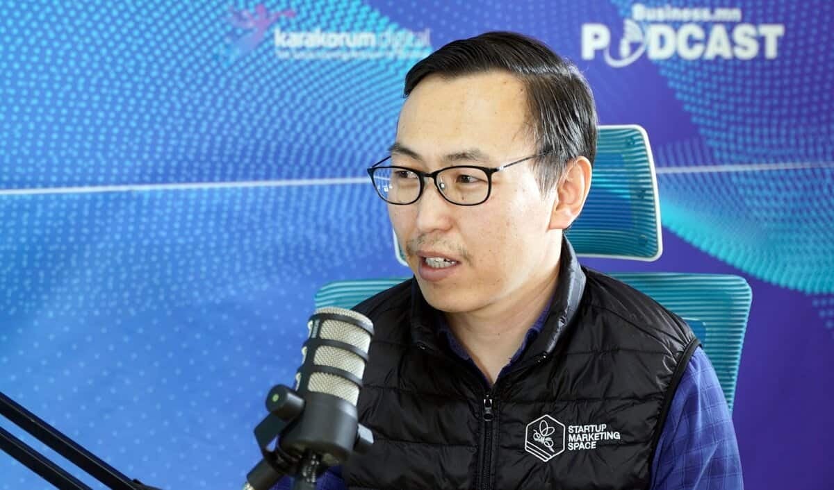 Нүдний шил, хар хантааз өмссөн эрэгтэй бизнесийн подкаст бичлэгийн үеэр микрофон руу ярьж байна. Арын дэвсгэр дээр "Business Podcast" гэсэн бичвэр бүхий цэнхэр өнгийн дизайн, зүүн дээд буланд лого нь янз бүрийн компани, энтрепренерүүдийн түүхийг онцолсон байна. - Business.MN