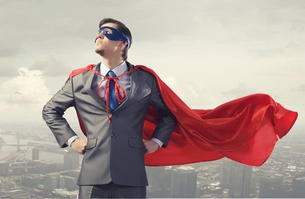 Улаан нөмрөг, цэнхэр маск өмссөн костюм өмссөн бизнес эрхлэгч, гараа ташаан дээрээ бариад итгэлтэйгээр зогсох, цаана нь хотын дүр төрх. - Business.MN