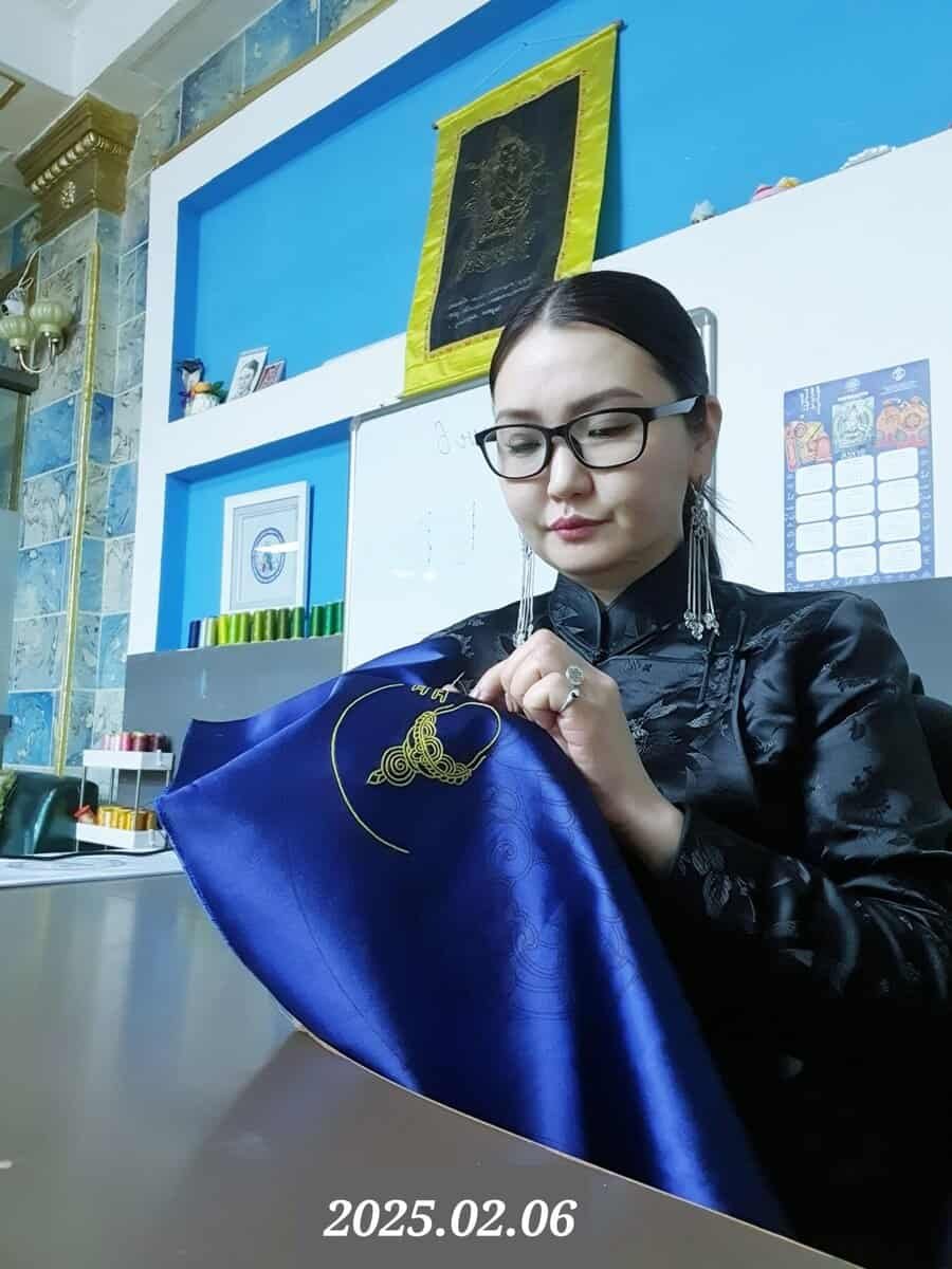 A person with glasses sits at a table, focusing on intricately stitching a gold thread design onto a piece of royal blue fabric. The background features a whiteboard, a calendar, and various decorations. The date "2025.02.06" is at the bottom.
