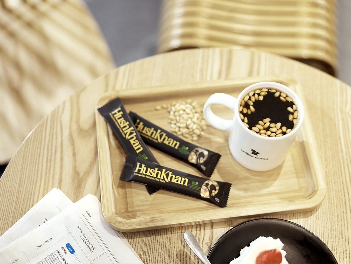 A wooden table with a tray holding a cup of coffee topped with pine nuts and two HushKhan snack bars. Nearby, there’s an open newspaper, a black plate with a dessert, and cutlery.