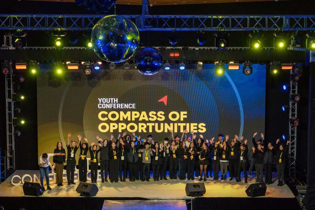 Албан ёсны хувцас өмссөн том бүлэг хүмүүс гараа өргөсөн тайзан дээр зогсож баяраа тэмдэглэж байна. Тэдний ард байрлах дэлгэцэн дээр "Залуучуудын бага хурлын боломжуудын луужин" гэсэн бичээсийг тод гэрэлтүүлж, таазнаас өлгөөтэй том диско бөмбөгнүүд нь залуу энтрепренерийн сүнсийг илэрхийлж байна. - Business.MN