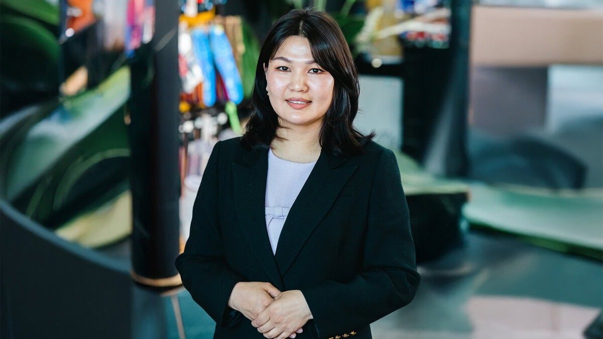 A woman with shoulder-length dark hair, wearing a black blazer and light-colored top, stands indoors with colorful blurred objects and modern decor in the background, smiling confidently like a model for Таныг хүндэтгэх гутлын брэнд.