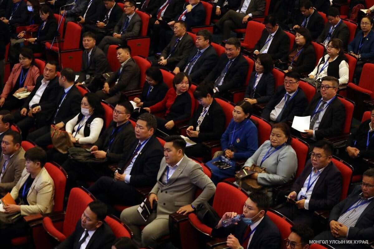 Албан ёсны хувцас өмссөн олон тооны хүмүүс улаан сандал дээр сууж, доторх арга хэмжээнд оролцож байна. Тэд анхааралтай урагшаа харж, зарим нь тэмдэглэл хөтөлж, зарим нь бичиг баримт барьдаг. Энэ нь бизнесийн хурал эсвэл компанийн удирдлагууд болон энтрепренерүүдийн албан ёсны уулзалт юм шиг санагдаж байна. - Business.MN