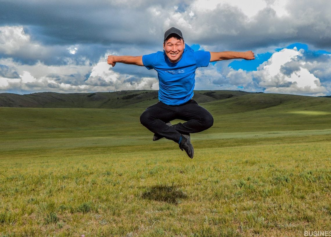 Цэнхэр цамц, хар малгайтай хүн гар, хөлөө өргөн агаарт баяр хөөртэй үсэрч байна. Тэд бага зэрэг үүлтэй тэнгэрийн дор уудам өвслөг талбайд гадаа байна. - Business.MN