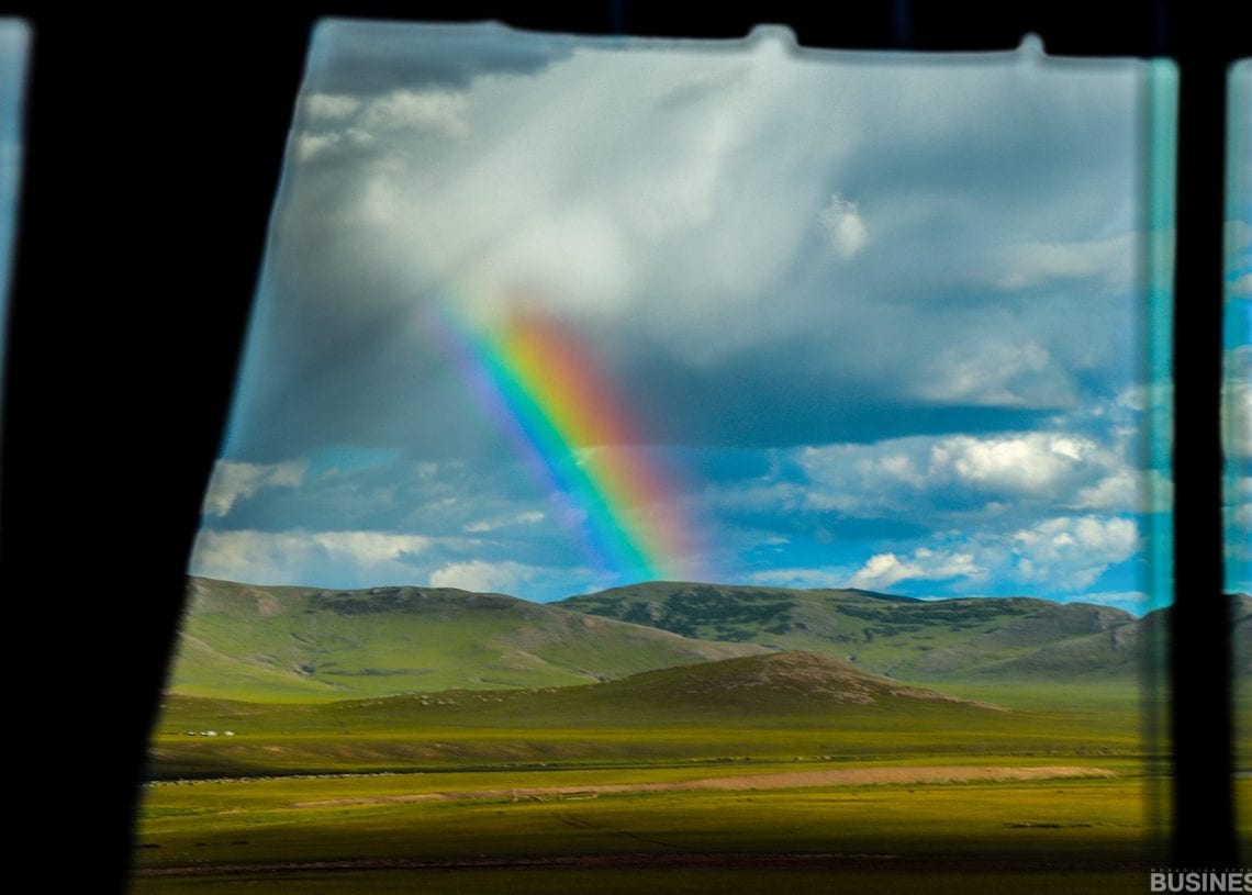 Цонхоор авсан уудам ногоон ландшафт дээрх эрч хүчтэй солонгын үзэмж. Тэнгэр бага зэрэг үүлэрхэг, алс холын талбай, толгод дээр сүүдэрлэж байна. - Business.MN