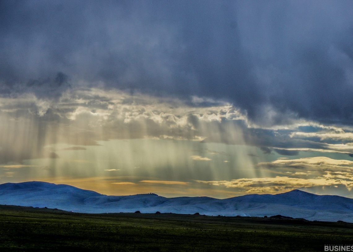 Өтгөн үүлийг нэвтлэн нарны туяа туссан гайхалтай тэнгэрийн доорх цаст уулс. Урд тал нь өргөн уудам, тэгш ногоон талбайг харуулдаг бөгөөд энэ нь хар үүл болон гэрэлтсэн ландшафтын хооронд эрс ялгаатай байдлыг бий болгодог. - Business.MN