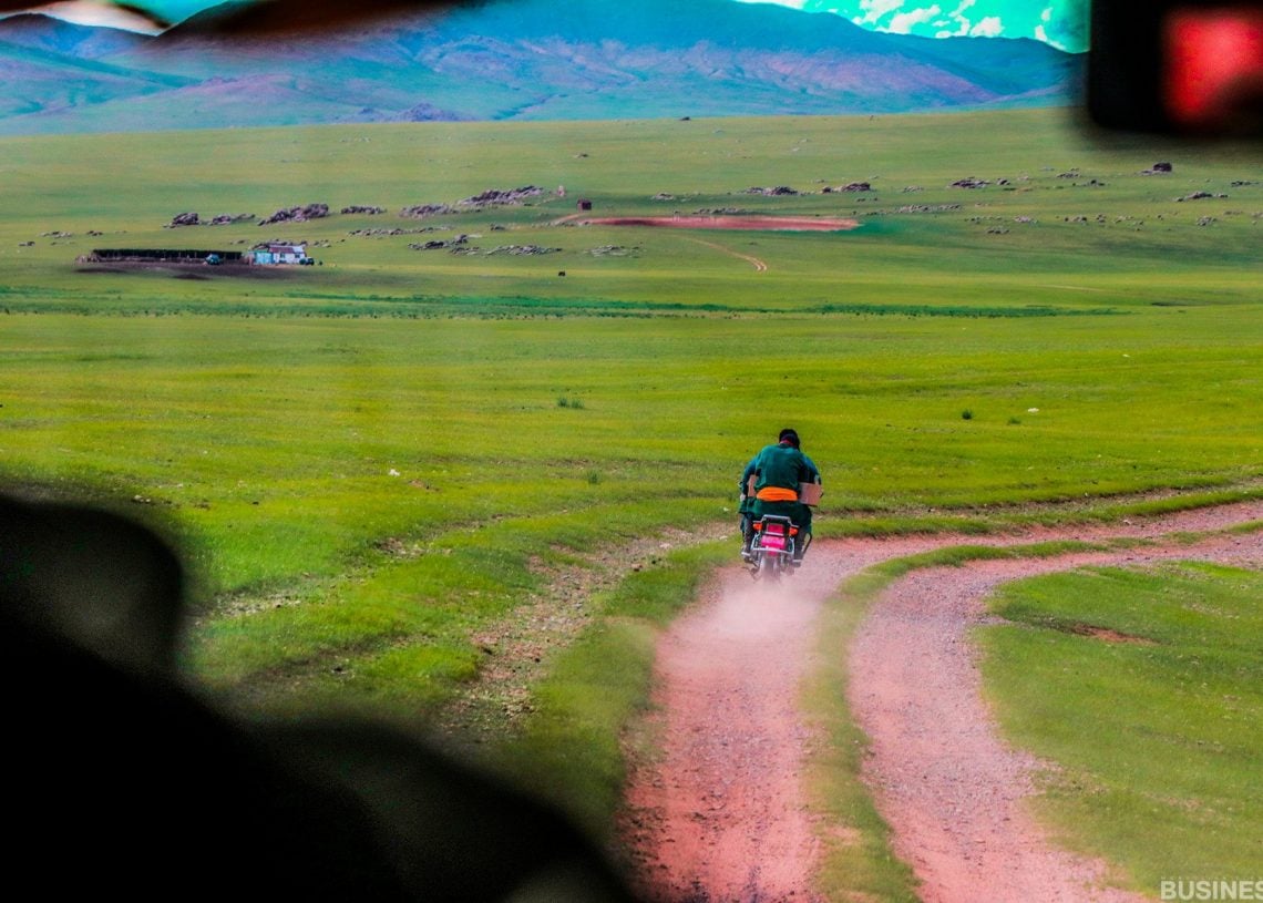 Өргөн уудам ногоон зүлэгт шороон замаар мотоцикль унаж яваа хүн. Үүлэрхэг дов толгод, алсын мал сүрэг үүлэрхэг тэнгэрийн дор харагдана. Тээврийн хэрэгслийн салоны хэсэг нь үзэмжийг бүрдүүлдэг. - Business.MN