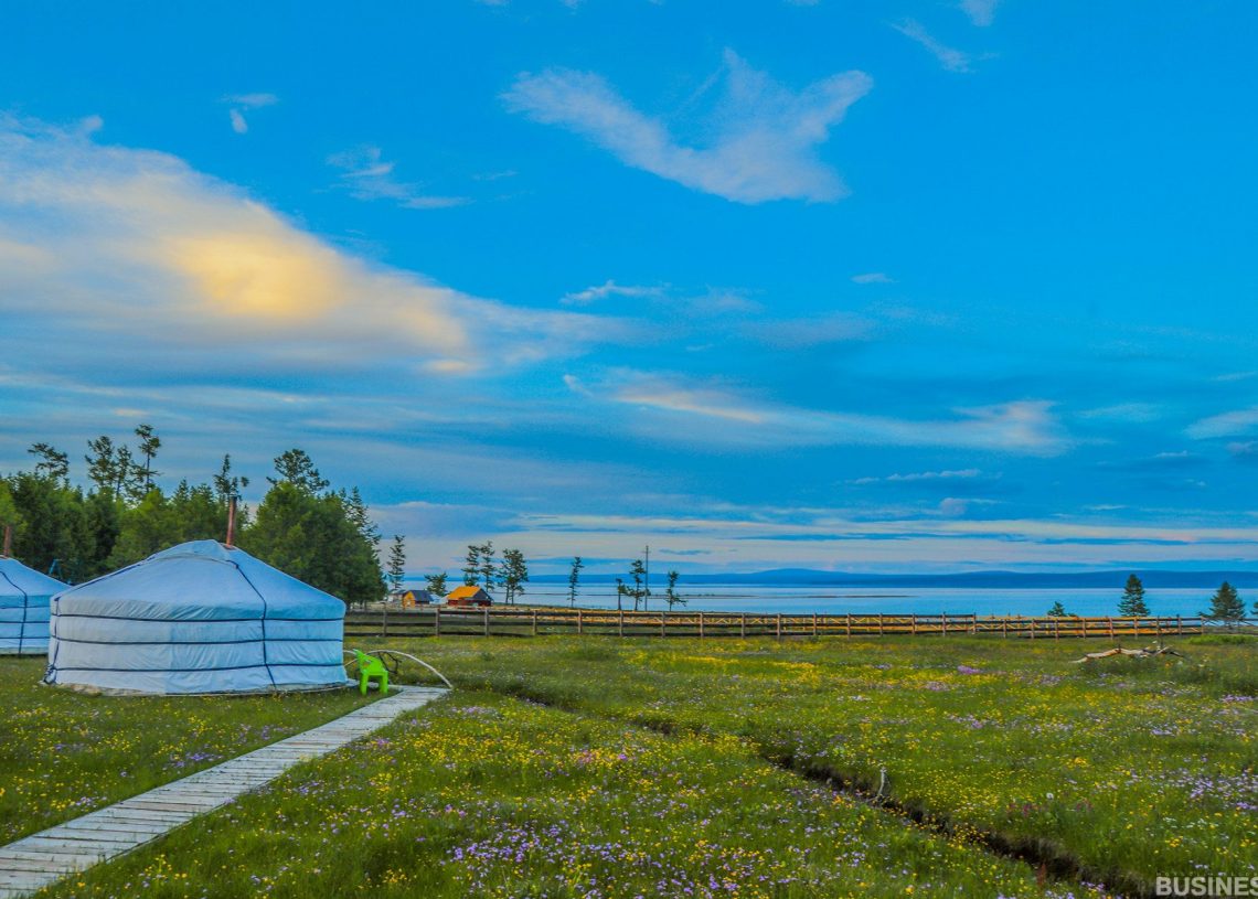 Цэлмэг хөх тэнгэрийн дор хоёр өргөөтэй өвслөг талбайн үзэсгэлэнт төрх. Модон зам нь гэрийн байшин руу хөтөлдөг. Цаана нь модон хашаа, модоор хүрээлэгдсэн алс холын усан сан харагдана. - Business.MN