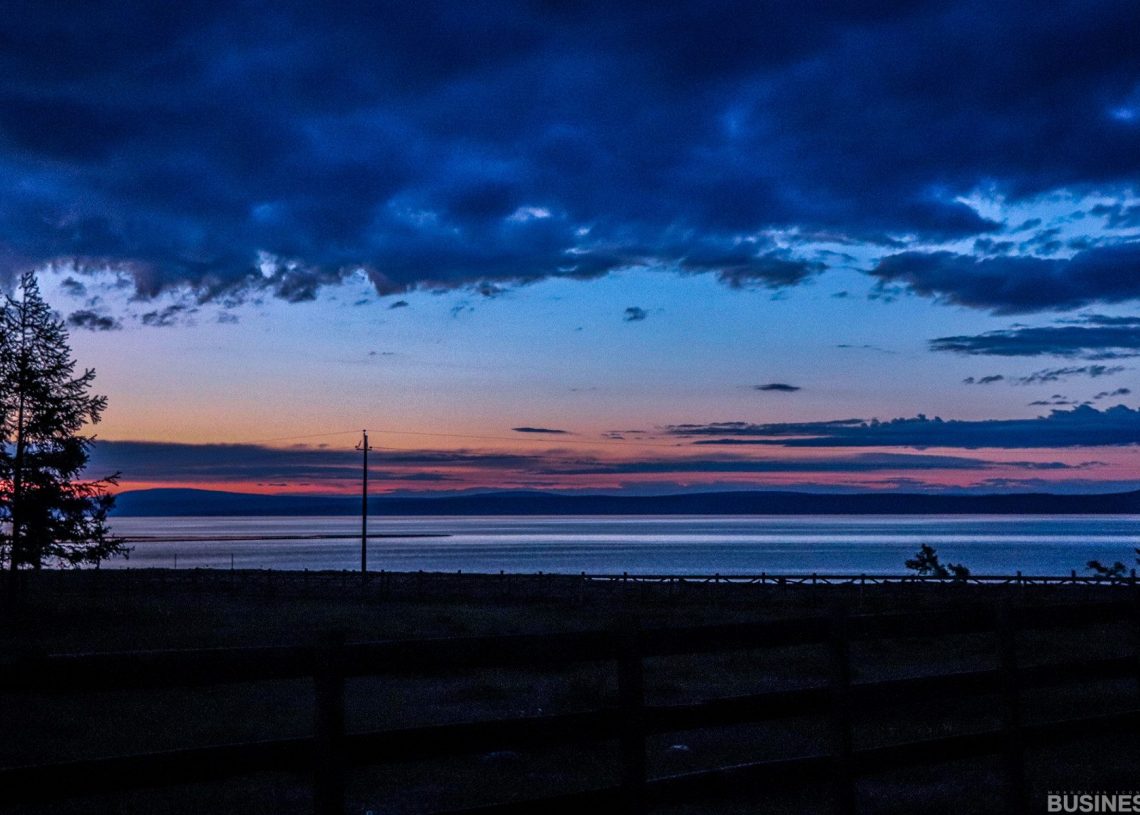 Тэнгэрт улбар шараас гүн цэнхэр хүртэл эрч хүчтэй градиент бүхий нуурын дээгүүр нам гүмхэн харанхуй дүр зураг. Урд талд нь мод, хашааны дүрс харагдана, харин цөөхөн тооны үүлс нь үдшийн амгалан тайван уур амьсгалыг сайжруулна. - Business.MN
