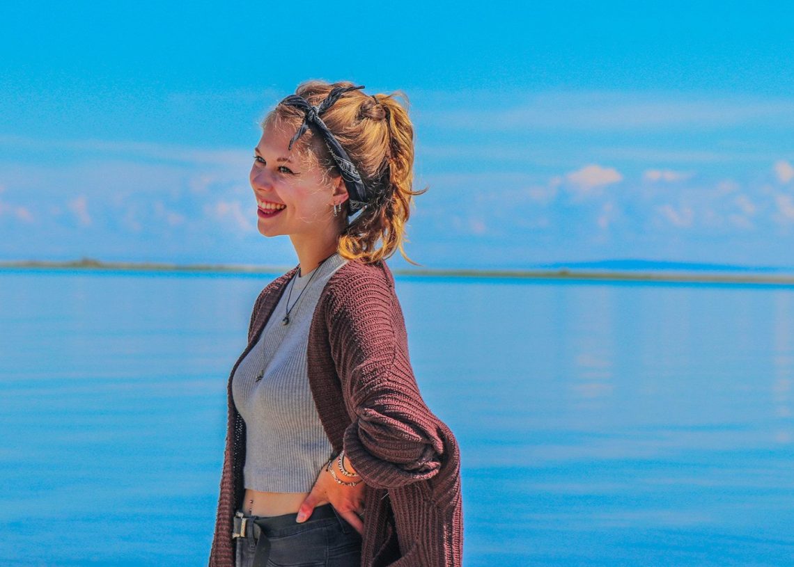 Толгойн боолттой инээмсэглэсэн хүн хөх тэнгэрийн дор тайван нуурын дэргэд зогсож байна. Тэд хүрэн өнгийн кардиган, саарал цамц өмсөж, нэг гараа ташаан дээрээ тавьж, тайван орчинг таашааж байна. - Business.MN