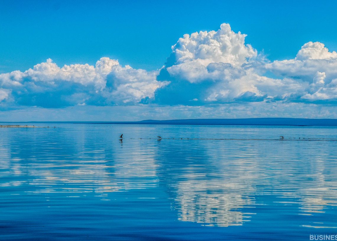Хурц хөх тэнгэр, сэвсгэр цагаан үүлсийг тусгасан намуухан нуурын намуухан дүр зураг. Алсын зайд шувуудын цуваа усны гадаргуу дээгүүр гүйлдэнэ. Тэнгэрийн хаяа нь алс холын газрын бүдэгхэн тоймоор хиллэдэг. - Business.MN