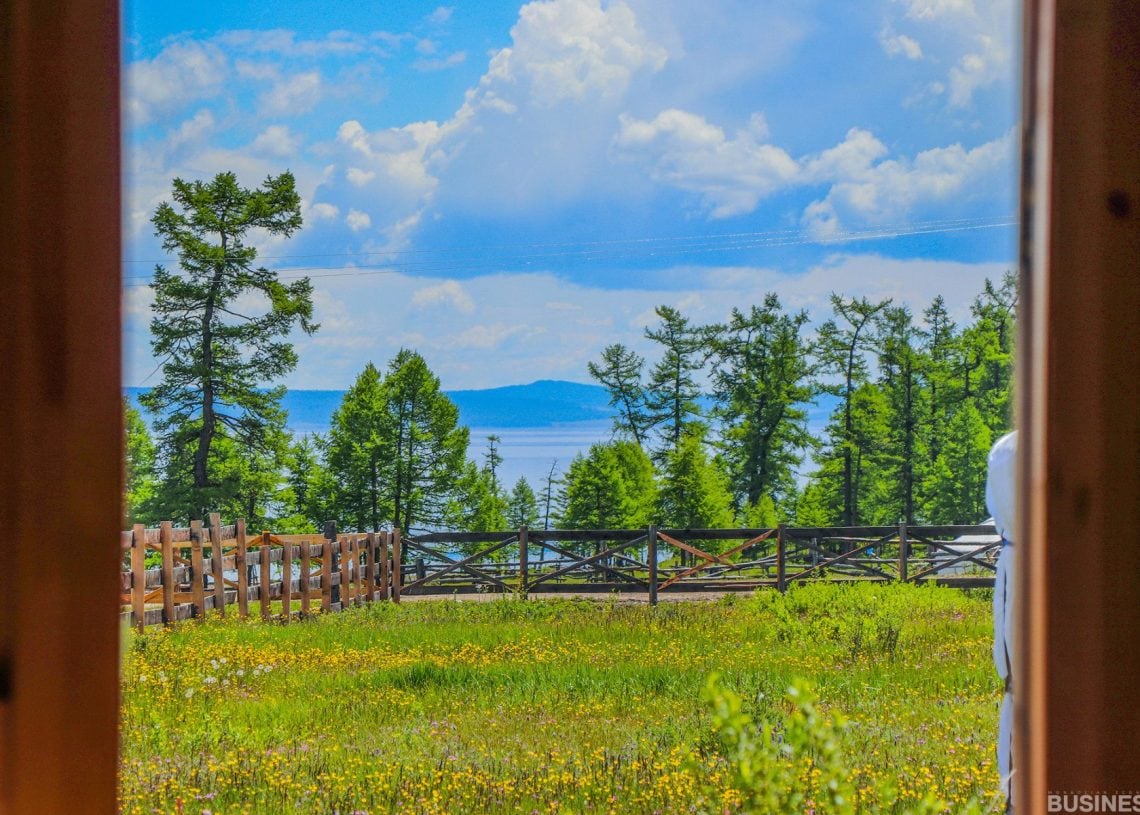 Онгорхой хаалгаар харвал шар цэцэг, модон хашаа, нарс мод бүхий өвслөг талбайг харна. Багавтар үүлтэй тэнгэрийн дор нуур, уулс харагдана. - Business.MN