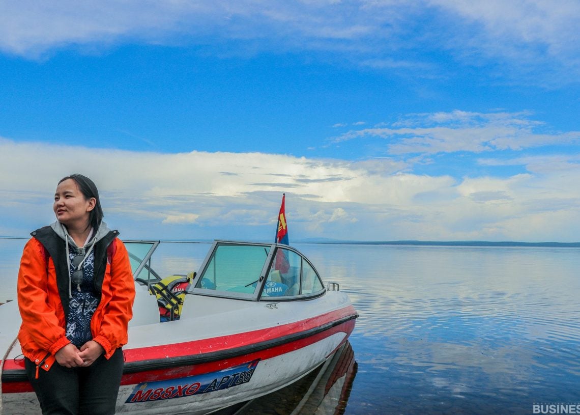 Улбар шар өнгийн хүрэм өмссөн хүн "Maska A320" гэсэн шошготой цагаан завины дэргэд, үүл бүрхсэн тод цэнхэр тэнгэрийн дор тайван, уудам нуурын дэргэд зогсож байна. Усан онгоцон дээр туг байх бөгөөд цаана нь тэнгэрийн хаяа сунадаг. - Business.MN