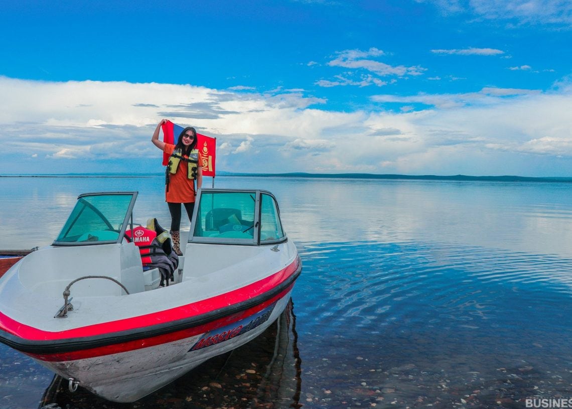 Аврах хантааз өмссөн хүн нам гүм нууран дээр жижиг завин дээр зургаа авахуулж байна. Тэнгэр нь сарнисан үүлстэй хөх өнгөтэй, ус нь тэнгэрийг тусгадаг. - Business.MN
