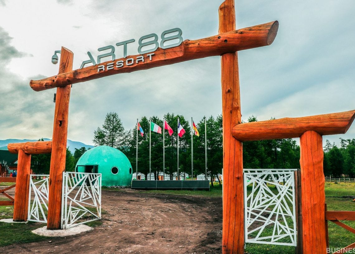 Том модон нуман хаалгатай Art88 амралтын газрын үүд. Хаалга нь бөмбөгөр хэлбэртэй барилга, шон дээр хэд хэдэн туг далбаа руу хөтөлдөг. Цаана нь мод, бүрхэг тэнгэр харагдана. - Business.MN