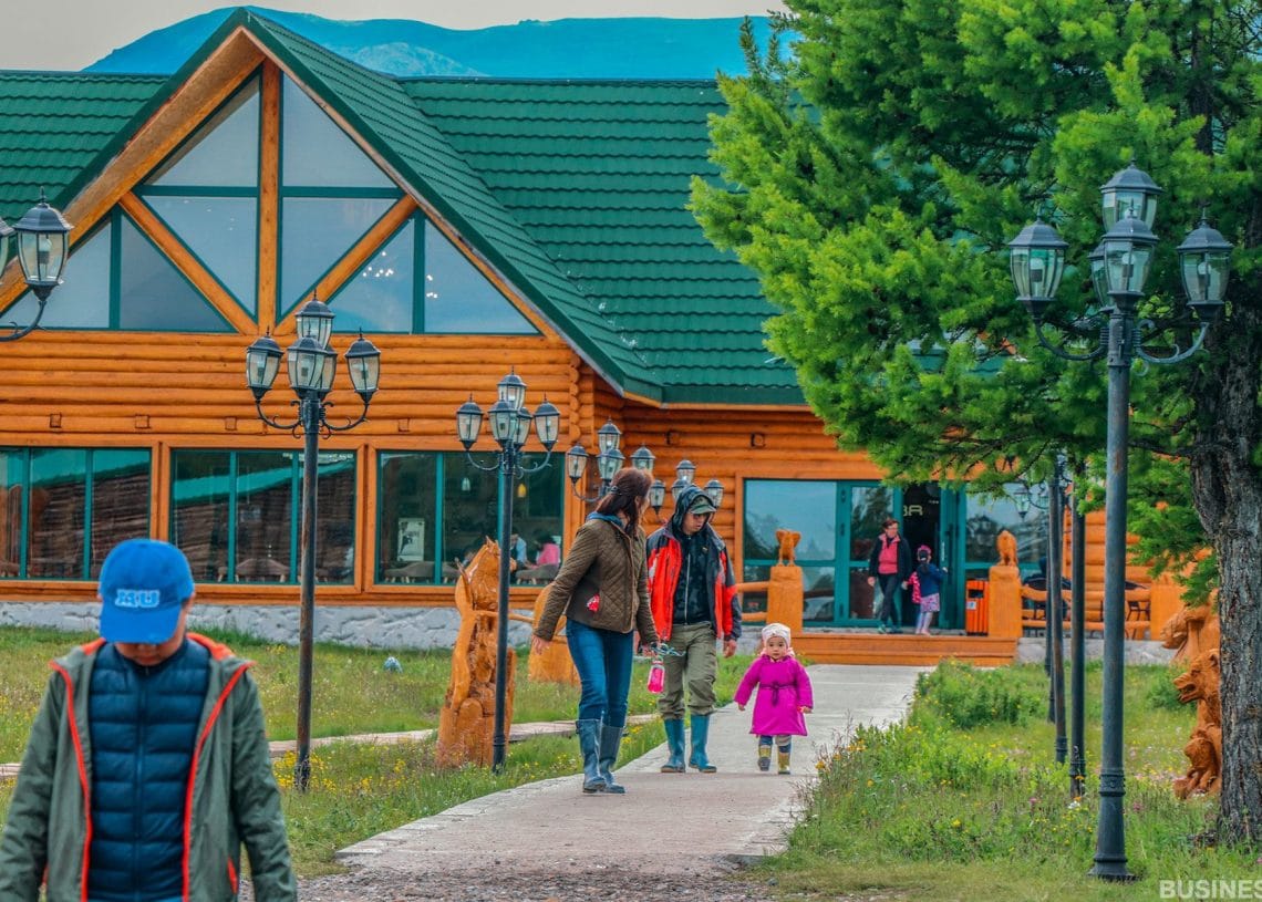 Нэг гэр бүл мод, гэрлийн шонгоор хүрээлэгдсэн ногоон дээвэр бүхий дүнзэн байшин руу чиглэн алхаж байна. Ягаан хувцастай хүүхэд насанд хүрэгчидтэй хөтлөлцөж байхад цэнхэр малгайтай хүү урагш алхаж байна. Арын дэвсгэр нь уулс, цэлмэг тэнгэрийг харуулдаг. - Business.MN