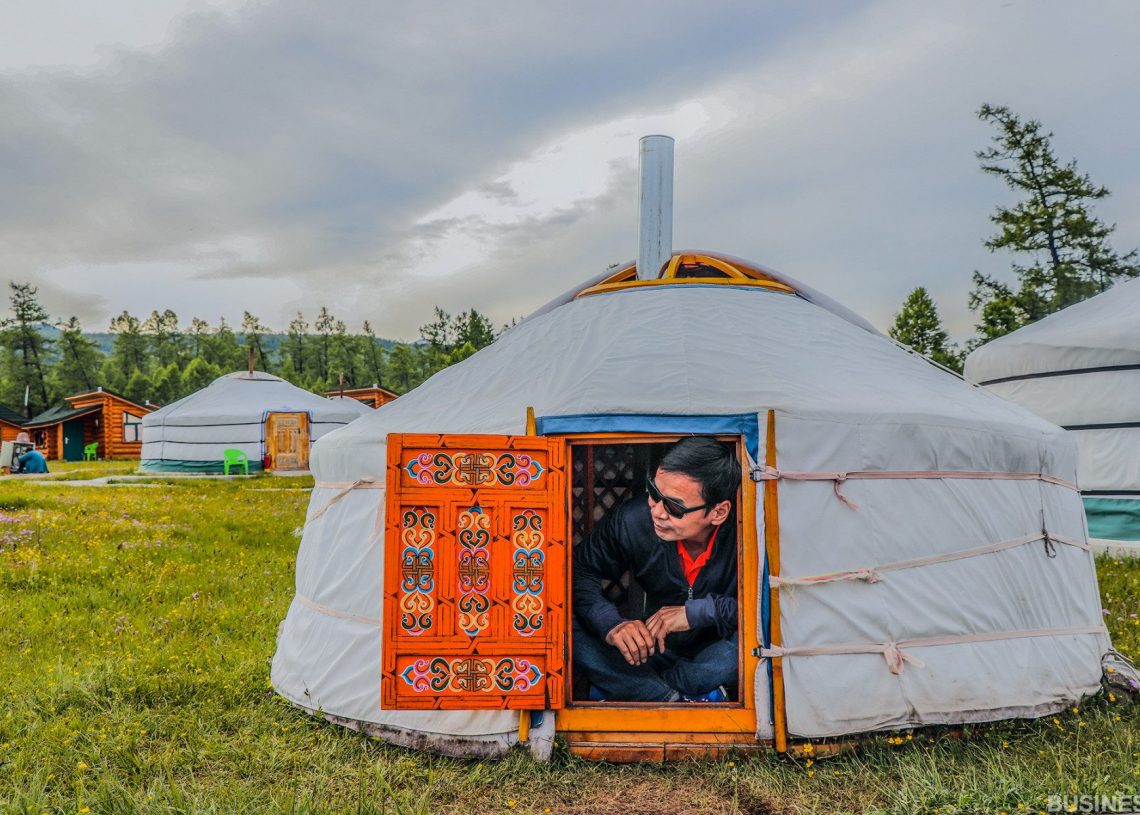 Нарны шил зүүсэн хүн гоёл чимэглэлтэй модон хаалгатай үндэсний гэрийн үүдэнд бөхийж байна. Юрт нь бүрхэг тэнгэрийн дор, мод, нэмэлт гэрүүдээр хүрээлэгдсэн өвслөг талбайд байрладаг. - Business.MN