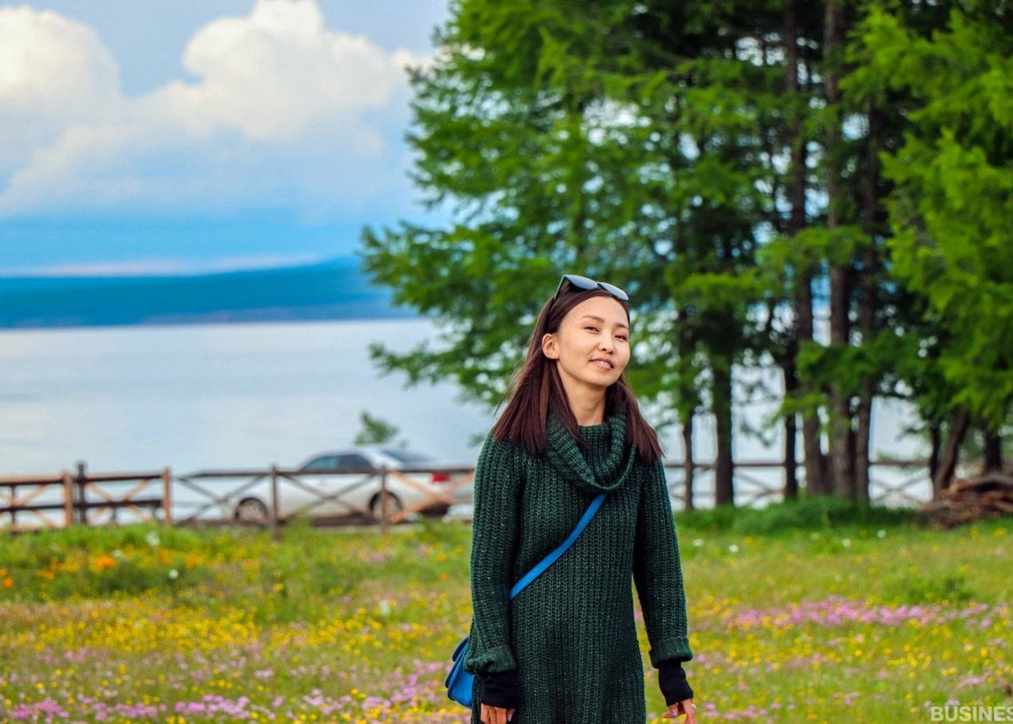 Өнгө өнгийн зэрлэг цэцэгсээр дүүрсэн өвслөг хээр дундуур инээмсэглэн алхаж буй эмэгтэй. Тэр хар ногоон цамц өмсөж, толгой дээрээ нарны шил зүүсэн байна. Түүний цаана бага зэрэг үүлэрхэг тэнгэртэй нуур, мод харагдана. - Business.MN