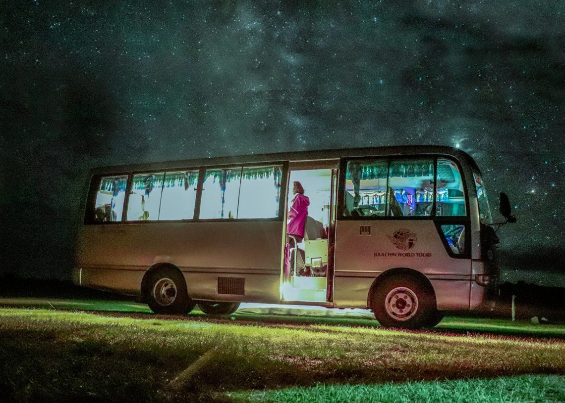 Шөнийн цагаар гэрэлтдэг автобус, одтой тэнгэрийн дор өвслөг талбай дээр зогсож байв. Дотор нь гэрэлтэй, орцны дэргэд хүн харагдана. Гадна тал нь "БИЗНЕС" гэсэн бичвэр, одод нь тайван уур амьсгалыг нэмдэг. - Business.MN