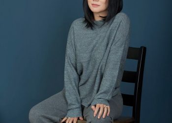 Хар үстэй хүн саарал өнгийн ноолууран хувцас өмсөж, шаргал өсгийтэй гутал өмссөн цэнхэр хананд сандал дээр сууна. Зүүн дээд буланд "Ноолуурын цех" гэсэн бичвэр байна. - Business.MN