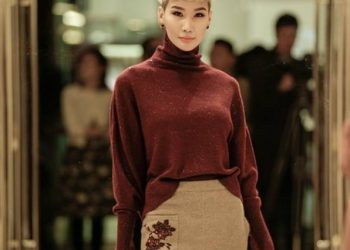 Богино цайвар үстэй хүн хүрэн улаан цамцтай, цэцэгсийн хатгамалтай шаргал банзал өмссөн байна. Тэд гэрэлтүүлэг сайтай өрөөнд дээр нь лааны суурь тавьж, ард нь бүдэг бадаг хүмүүстэй итгэлтэйгээр зургаа авахуулж байна. - Business.MN