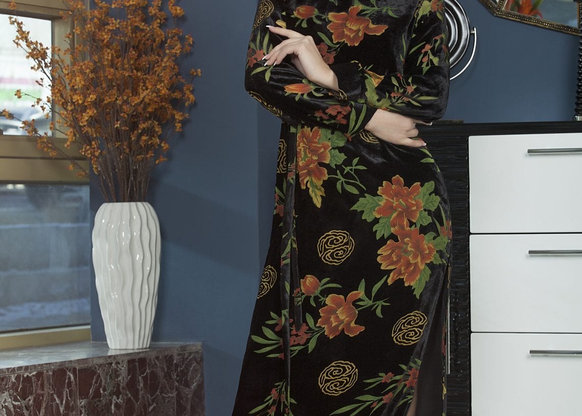 Өндөр ангархайтай урт хар цэцэгтэй даашинз өмссөн эмэгтэй гоёмсог өрөөнд инээмсэглэн зогсож байна. Тэр гоёл чимэглэлийн толь бүхий шүүгээний хажууд байх бөгөөд гантиг цонхны тавцан дээр улбар шар цэцэгтэй ваар байдаг. Текст "Хувцасны цех. - Business.MN