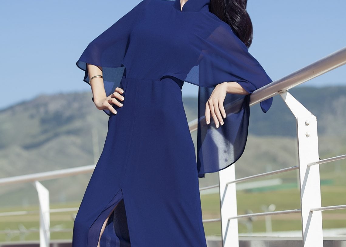 Цэнхэр даашинзтай эмэгтэй тавцан дээр дэгжин зургаа авахуулж байна. Тэр цэлмэг хөх тэнгэрийн дор хашлага налан, цаана нь толгод харагдаж байна. Тод бичвэрт зүүн дээд буланд "ХУВЦАСНЫ ҮЙЛЧИЛГЭЭ" гэж бичнэ. - Business.MN