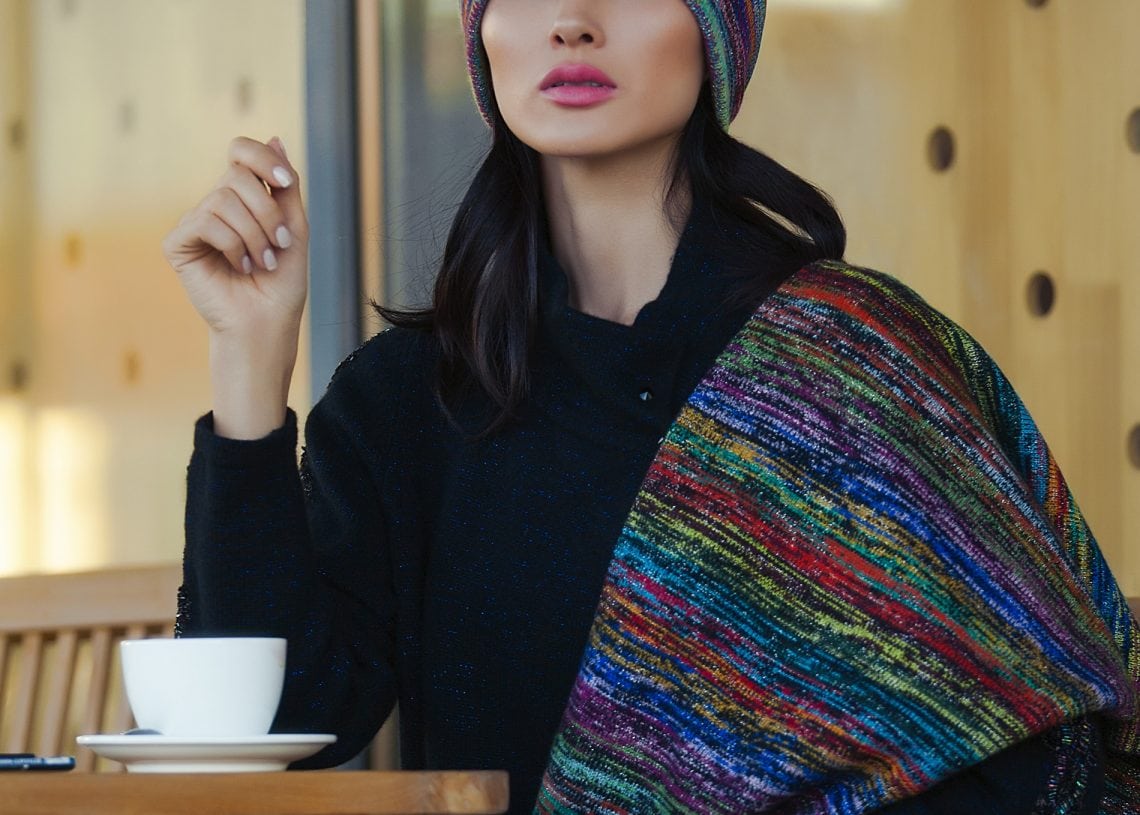 Өнгө өнгийн сүлжмэл малгай, алчуураа өмссөн хүн аяга кофе бариад модон ширээний ард сууна. Арын дэвсгэр нь дугуй зүсэлт бүхий модон хавтангаар хийгдсэн байдаг. Дээр "НООЛУУРЫН ЦЕХ" гэсэн бичиг харагдаж байна. - Business.MN