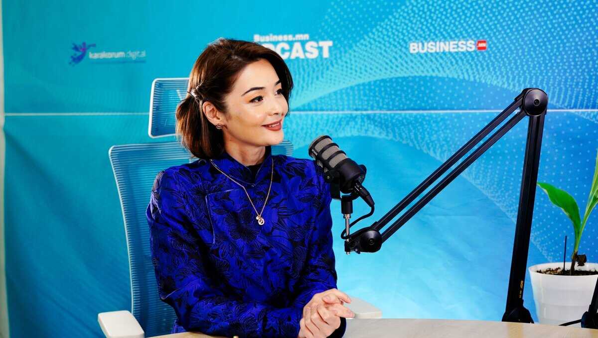 Цэнхэр хээтэй цамц өмссөн мөр хүртэл урт үстэй эмэгтэй ширээний ард сууж, тохируулж болох гараараа микрофоноор ярьж байна. Тэр аятайхан илэрхийлэлтэй бөгөөд арын дэвсгэр дээр бизнес подкасттай холбоотой текст, лого багтсан байна. - Business.MN