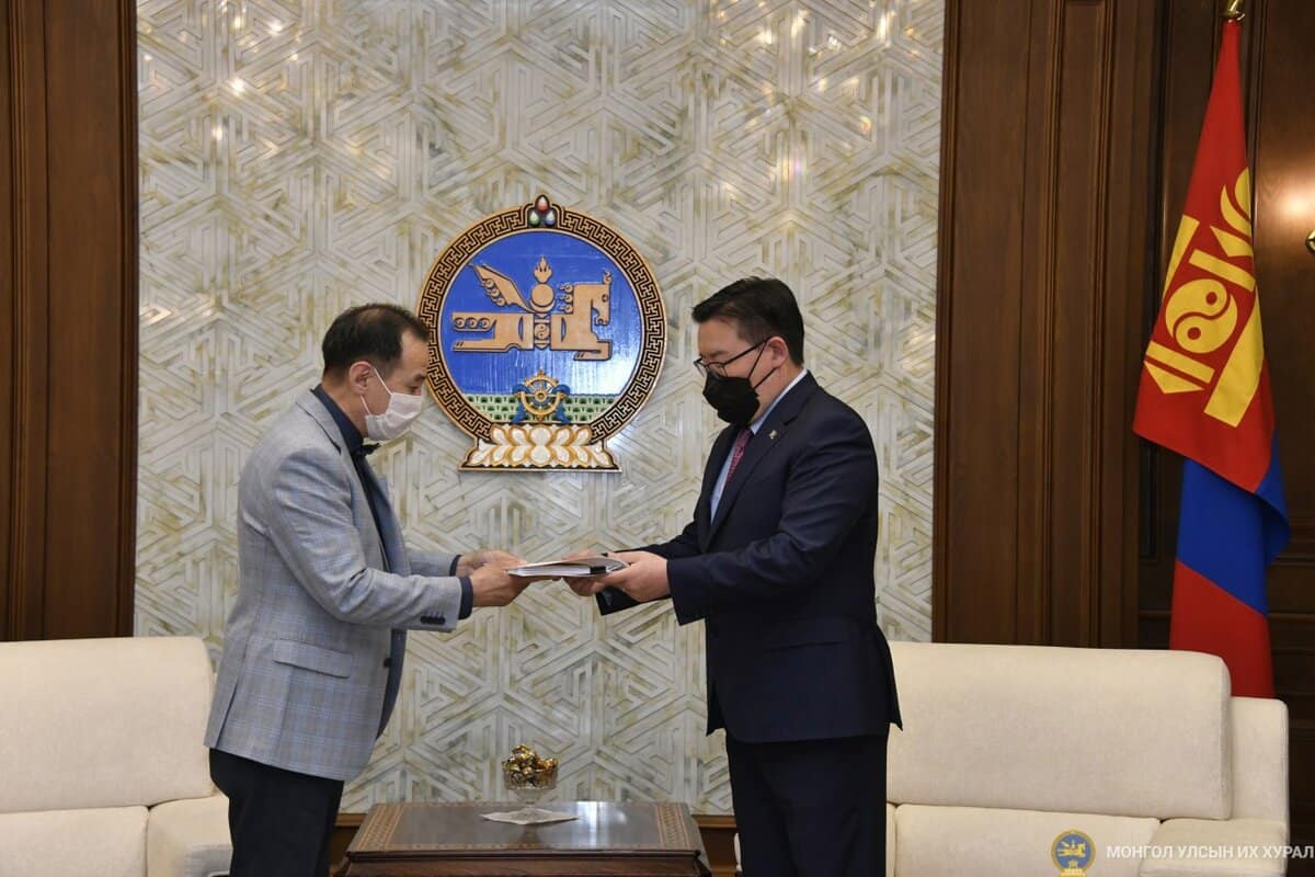 Нүүрний маск зүүсэн хоёр эрэгтэй баруун талд шар, улаан өнгийн туг бүхий том бэлгэ тэмдэг бүхий гоёл чимэглэлийн өрөөнд зогсож байна. Тэд бичиг баримтаа солилцож байна. Хоёулаа ажил хэргийн уулзалтад зориулагдсан костюм өмссөн байна. Зүүн талд байгаа хүн саарал костюмтай, баруун талд байгаа хүн бараан костюмтай. - Business.MN